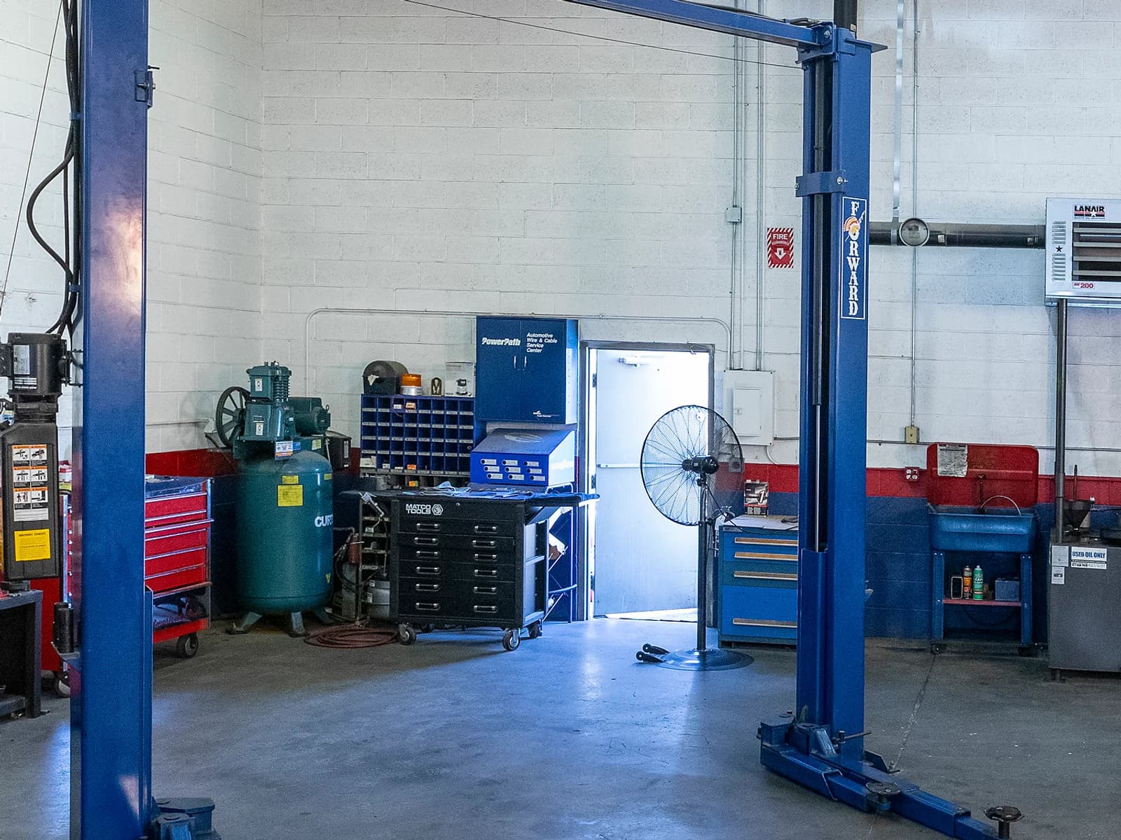 Air compressor, blue tool cabinet and a fan beside a lift post and an open door