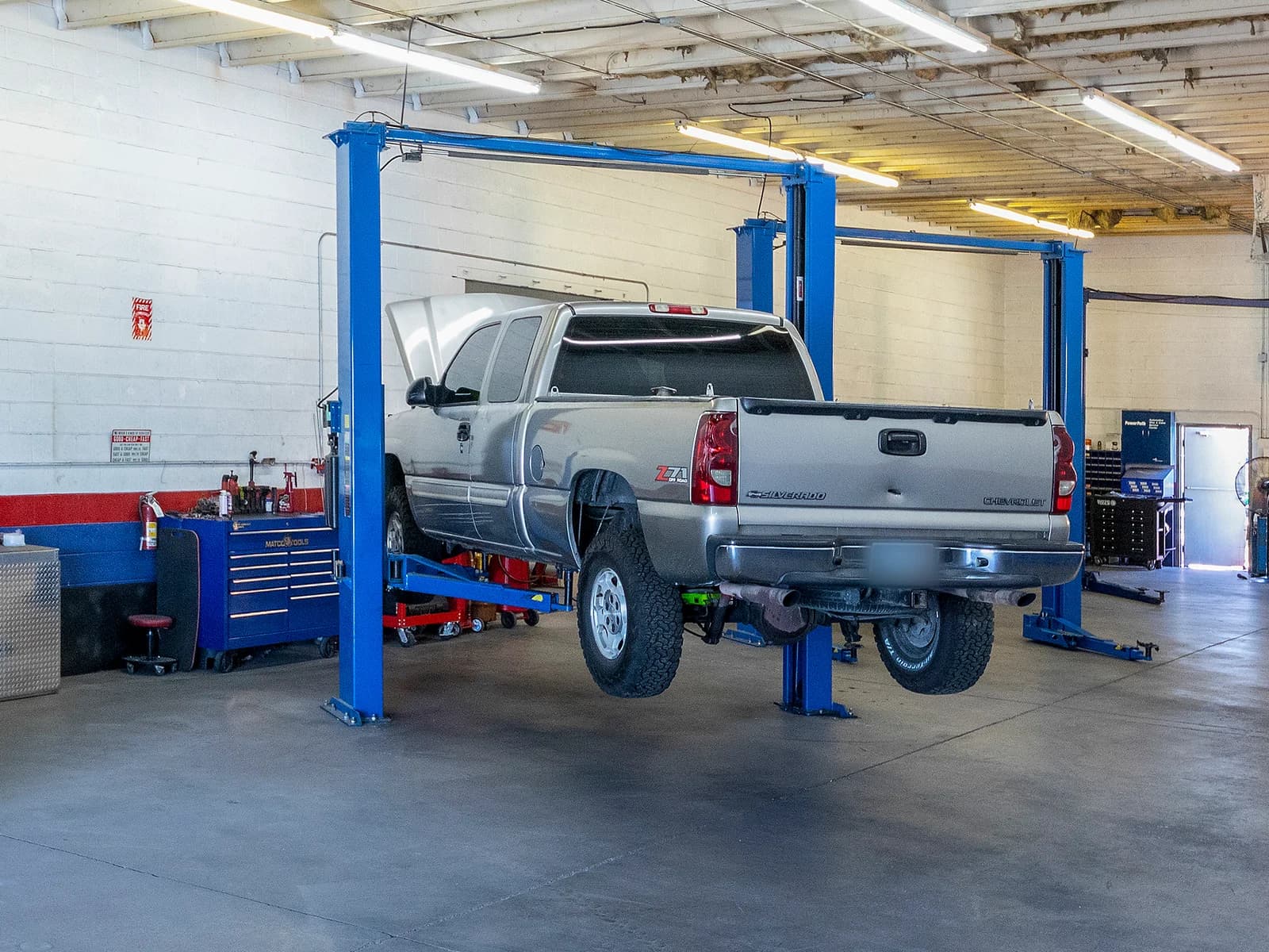 Silver work pickup with its hood open, raised on a lift in a bay